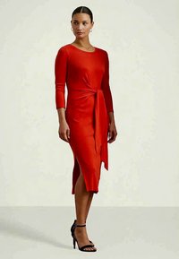 Γυναίκα που φοράει ένα εφαρμοστό κόκκινο φόρεμα midi με τρία τέταρτα μανίκια, πλαϊνό σκίσιμο και λεπτομέρεια με κομμένα κορδόνια στη μέση, που έχει συνδυαστεί με μαύρες ψηλοτάκουνες σάντερς.