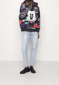 Sudadera con capucha colorida que presenta un gran dibujo de Mickey Mouse, fabricada con una tela suave y un ajuste holgado, combinada con pantalones vaqueros azul claro y mocasines negros.