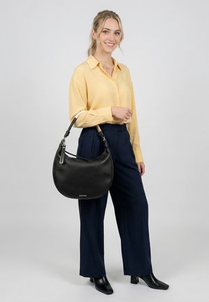 Frau in gelbem Hemd und dunkelblauer Hose hält eine schwarze Lederhandtasche mit einem Schulterriemen und lächelt vor einfarbigem Hintergrund.
