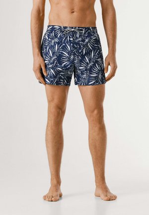 Mann trägt marineblaue Badeshorts mit weißem Blattmuster, steht barfuß vor einem schlichten hellen Hintergrund.