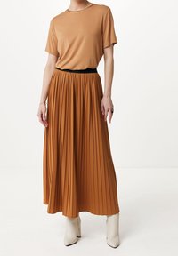 Falda marrón plisada con cintura elástica negra, combinada con una blusa marrón de manga corta. La modelo lleva botas blancas hasta el tobillo. Fondo neutro.