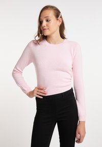 Maglione lavorato a maglia rosa chiaro con maniche lunghe e orlo a coste. La texture sembra morbida; abbinato a pantaloni neri per il contrasto.