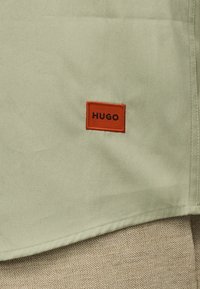 Ανοιχτοπράσινο ύφασμα με ένα μικρό κόκκινο ορθογώνιο πατς ραμμένο, που εμφανίζει τη λέξη «HUGO» με μαύρα κεφαλαία γράμματα.