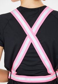 Musta paita, jossa on vaaleanpunaisia ja valkoisia heijastavia hihnoja ristiin selässä, ja joka omaa sileän pinnan sekä säädettävän solkipoman.