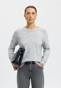 Világosszürke, gyapjúkeverék pulóver kerek nyakkal, magas derekú fekete farmerrel párosítva. A modell egy fekete bőr táskát tart, amely egyedi formájú.