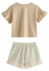 Beige kortærmet top med flæser ved ærmerne, parret med hvide shorts med grønt zigzag-mønster og blondekant i bunden.