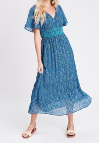 Robe longue à motifs bleus avec manches courtes volantées, taille smockée et jupe fluide. Présente un design géométrique avec des accents verts.