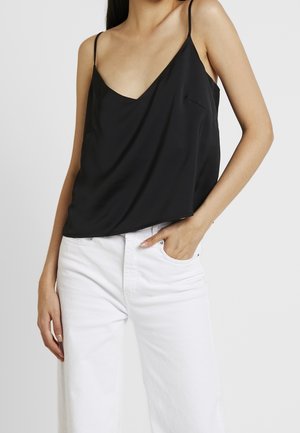 Femme portant un haut noir sans manches à encolure en V avec de fines bretelles et un pantalon blanc taille haute, une main dans la poche, sur un fond uni.