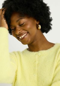 Cardigan jaune flou avec un col rond et une fermeture à cinq boutons, associé à des boucles d'oreilles en or. Tissu texturé à l'apparence douce.