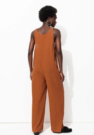 Combinaison sans manches, ample et marron, avec un tissu texturé, présentant des jambes larges et des fentes sur les côtés. Modèle portant de grandes boucles d'oreilles créoles.
