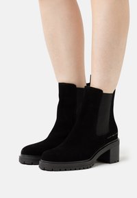 Botines de ante negro con tacón grueso, paneles laterales elásticos y un acento de logo en el tacón. Superficie texturizada con un acabado suave.