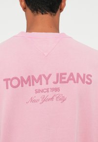 Gaiši rozā džemperis ar izteiksmīgu "TOMMY JEANS" logo, teksturēts audums, un v veida kakla izgriezums. Zem tā ir izdrukāts "Jaunā Jorka".