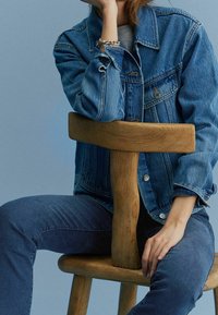 Chaqueta de mezclilla en azul claro, con bolsillos frontales y cierres de botones, combinada con jeans azul oscuro. Modelo sentado en una silla de madera.