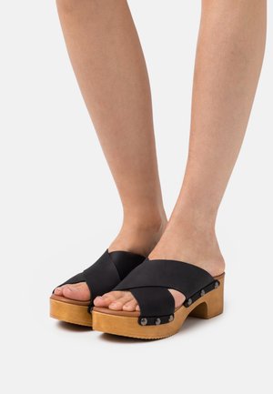 Mustat ristiolkapohjaiset kengät, joissa on vaaleanruskea, teksturoitu pohja. Ominaisuuksina pyöreä varvasosa ja koristeelliset metalliset niitit sivuilla.