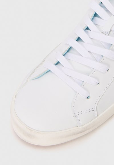 Baskets blanches en cuir avec des lacets blancs plats, une pointe arrondie, de subtils détails de couture et une surface lisse avec un bord de semelle légèrement usé.