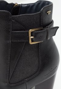Bottine noire avec un mélange de cuir et de tissu, un accent de boucle sur le côté et une finition texturée, avec un talon légèrement surélevé.