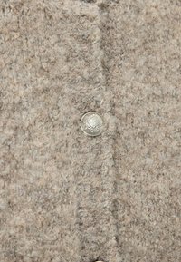 Brun tekstureret stof med en fuzzet overflade, der har en rund sølvknap med indviklet detaljering i midten.