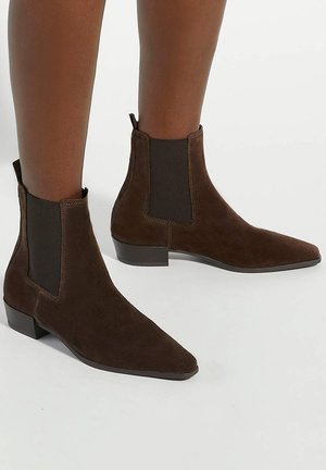 Bottines en daim marron avec panneaux latéraux élastiques et talons blocs bas portées sur des jambes à la peau foncée sur un fond uni.