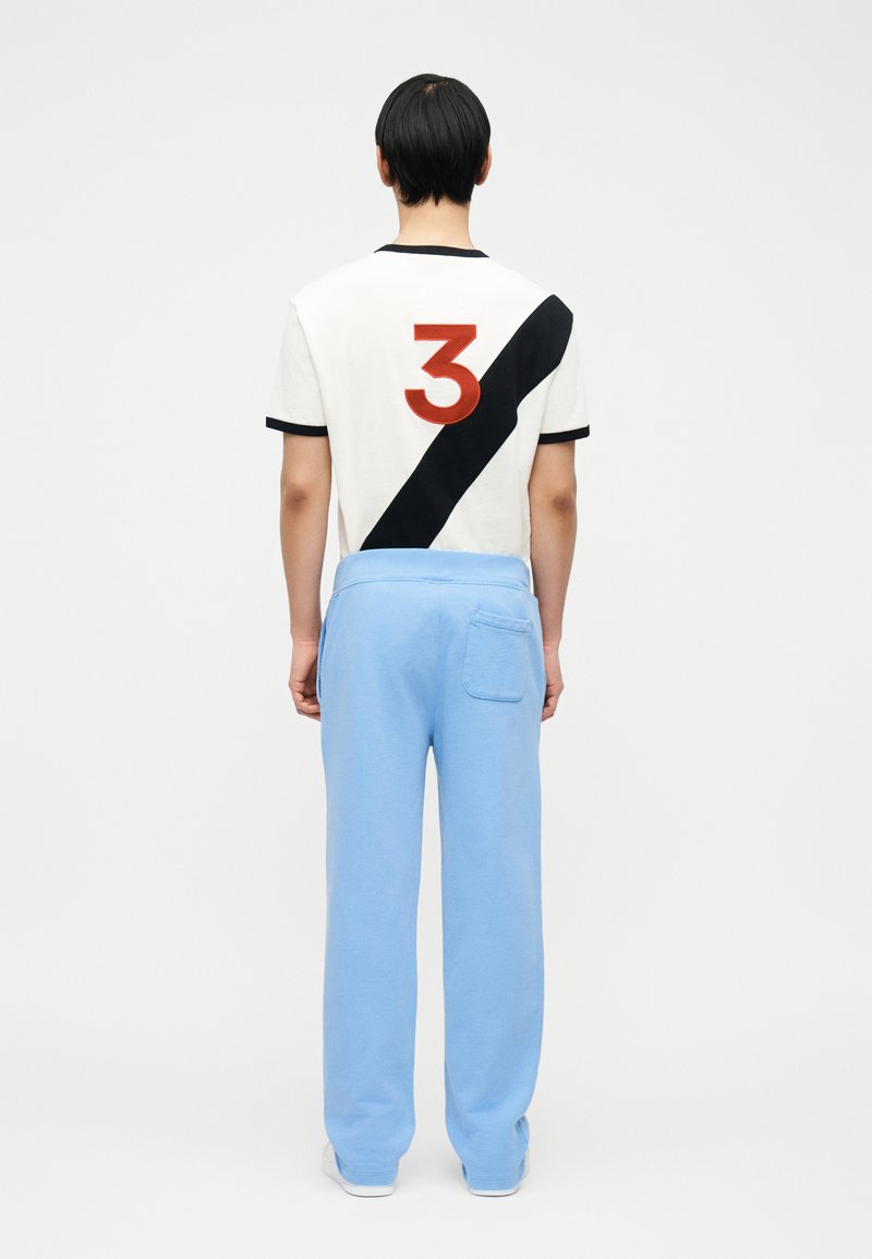 Personne debout, vue de dos, portant un t-shirt blanc avec une bande diagonale noire et le numéro 3 en rouge, associé à un pantalon bleu clair.