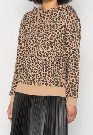 Frau trägt eine beige Leopardenmuster-Kapuzenjacke mit Kordeln und einen schwarzen Faltenrock, steht vor einem einfarbigen hellen Hintergrund.