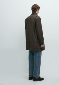 Brauner Mantel mit langen Ärmeln und geradem Schnitt, mit hohem Kragen und ohne sichtbare Verschlüsse, getragen über locker sitzende blaue Jeans aus Denim.