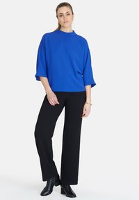 Blå, lös passande topp med hög krage, ihop med svarta, raka byxor och svarta ankelstövlar. Slät textur, modern design.