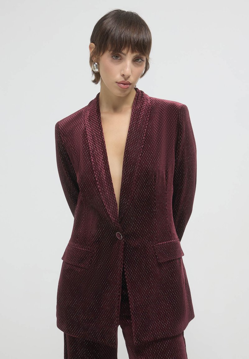 Blazer in velluto borgogna con scollatura a V profonda, due tasche frontali e chiusura a un bottone; superficie testurizzata con motivi sottili.