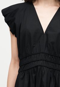 Γυναίκα που φοράει μαύρο φόρεμα με βαθύ V-λαιμόκοψη, φουσκωτά κοντά μανίκια και τρεις μαζεμένες ελαστικές ζώνες στη μέση.