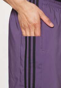 Fialové sportovní šortky s kapsou, mají černé pruhy podél strany. Hladký povrch a elastický pas pro pohodlí.