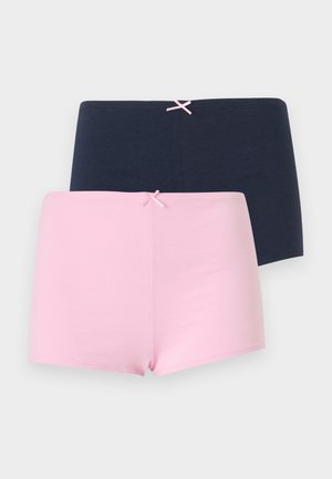 Jeune femme aux longs cheveux ondulés portant un caraco blanc avec des cœurs roses et un short rose, debout les mains sur les hanches.