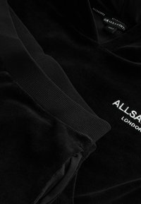 Juodas veliūro džemperis su raukiniais rankovėse, turintis V formos iškirpę ir išsiuvinėtą "ALLSAINTS LONDON" logotipą priekyje. Bendras minkštas tekstūra.