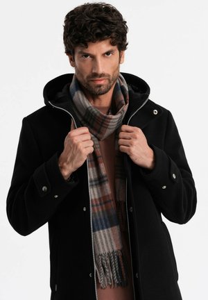 Black jacket with a hood, zip closure, and silver snaps. Worn with a plaid scarf featuring blue, brown, and beige tones.