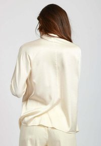 Blusa in seta crema con vestibilità rilassata, maniche lunghe, dettaglio della piega sul retro e orlo leggermente arrotondato. Texture liscia e aspetto leggero.
