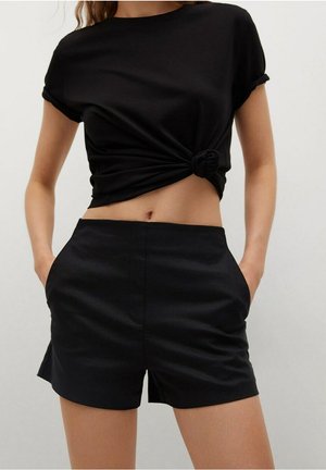 Donna che indossa un top corto annodato nero a maniche corte con risvolti e pantaloncini sartoriali neri a vita alta con tasche, in piedi con le mani in tasca.