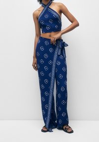 Navy sininen halterneck-crop top, jossa on valkoisia geometrisia kuvioita; yhteensopivat pitkät, ympärille kietoutuvat housut, joissa on solkityyli ja koristeellinen trimmi.