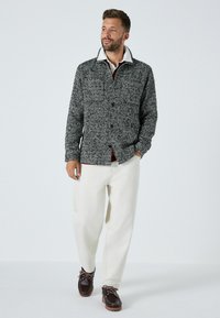 Sako s gombíkmi v čiernobielej houndstooth vzore, s preklápacím golierom, nosené cez marhuľovú košeľu, v kombinácii s krémovými nohavicami a tmavými topánkami.