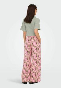 Femme aux longs cheveux foncés portant un haut vert sauge à manches courtes et un pantalon large rose à motifs, debout les mains dans les poches, de dos.