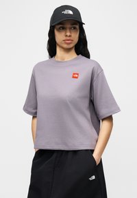 Γυναίκα που φοράει ένα λιλά κοντό μπλουζάκι, μαύρη φούστα και μαύρο καπέλο, όλα με το λογότυπο The North Face, στέκεται με τα χέρια στις τσέπες.