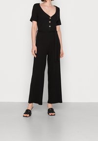 Czarny prążkowany kombinezon z krótkimi rękawami, dekoltem w kształcie litery V, pięcioma guzikami, wiązanym paskiem w talii oraz szerokimi spodniami. Połączony z czarnymi plecionymi klapkami.