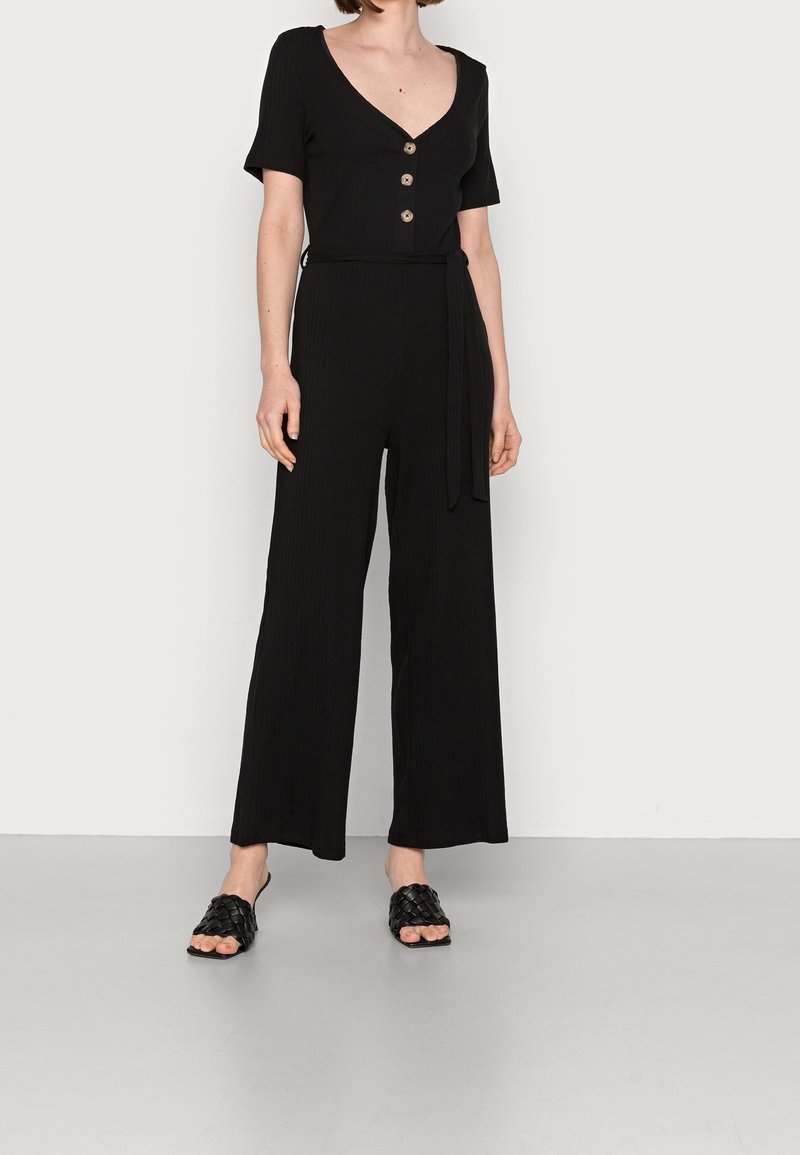 Czarny prążkowany kombinezon z krótkimi rękawami, dekoltem w kształcie litery V, pięcioma guzikami, wiązanym paskiem w talii oraz szerokimi spodniami. Połączony z czarnymi plecionymi klapkami.
