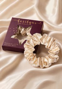 Beżowa jedwabna gumka do włosów obok ciemnofioletowego opakowania z okienkiem w kształcie gwiazdy i złotym napisem "lesilque luxury silk scrunchie" na beżowej satynowej tkaninie.