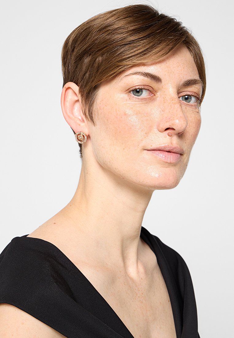 Frau mit kurzem braunem Haar, hellblauen Augen, die roségoldene Ohrringe und ein schwarzes ärmelloses Oberteil trägt und leicht nach rechts schaut.