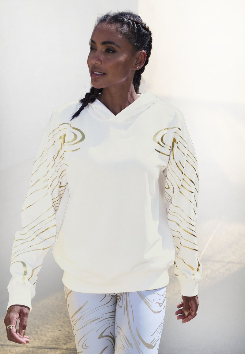 Cremfarbene Kapuzenjacke mit goldenen abstrakten Designs auf den Ärmeln, aus weichem Stoff gefertigt. Kombiniert mit leichten, gemusterten Leggings, glatte Textur.