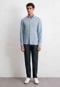 Lichtblauw langemouwen shirt met een button-down kraag, gecombineerd met donkere marineblauwe broek en witte sneakers, tegen een neutrale achtergrond.