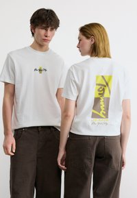 Δύο άτομα φορούν λευκά μπλουζάκια με γραφικά και σκούρα καφέ παντελόνια, το ένα κοιτά προς τα εμπρός και το άλλο δείχνει ένα μεγάλο σχέδιο στην πλάτη.