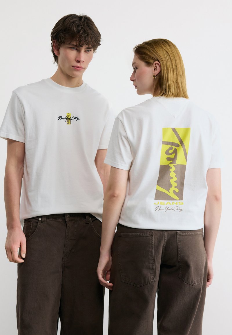 Δύο άτομα φορούν λευκά μπλουζάκια με γραφικά και σκούρα καφέ παντελόνια, το ένα κοιτά προς τα εμπρός και το άλλο δείχνει ένα μεγάλο σχέδιο στην πλάτη.