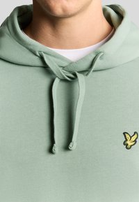 Felpa verde menta con cappuccio e cordoncino, in tessuto testurizzato, caratterizzata da un piccolo logo di un uccello nero e giallo sul davanti.