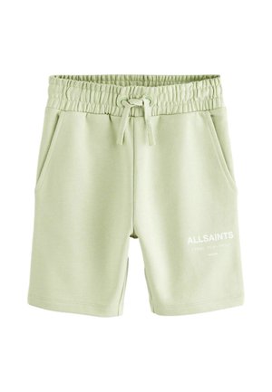 Jongen die een wit oversized T-shirt en lichtgroene korte broek draagt, met witte sokken en witte sneakers, staand tegen een effen grijze achtergrond.