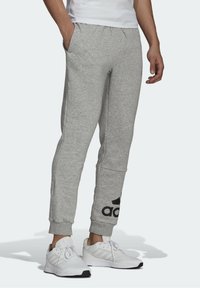 Szare spodnie dresowe o zwężanym kroju, z elastycznym paskiem w talii i kieszeniami. Na lewej nogawce znajduje się czarne logo Adidas. Materiał z mieszanki bawełny.