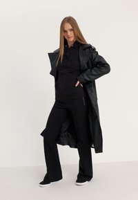 Svart kunstskinn lang jakke over en svart hettegenser, kombinert med svarte bukser og mørke sko. Outfitten har en avslappet passform og minimale detaljer.